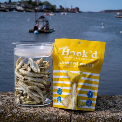 Jar of dried fish and 'Hook'd' package by a waterfront with boats in the background