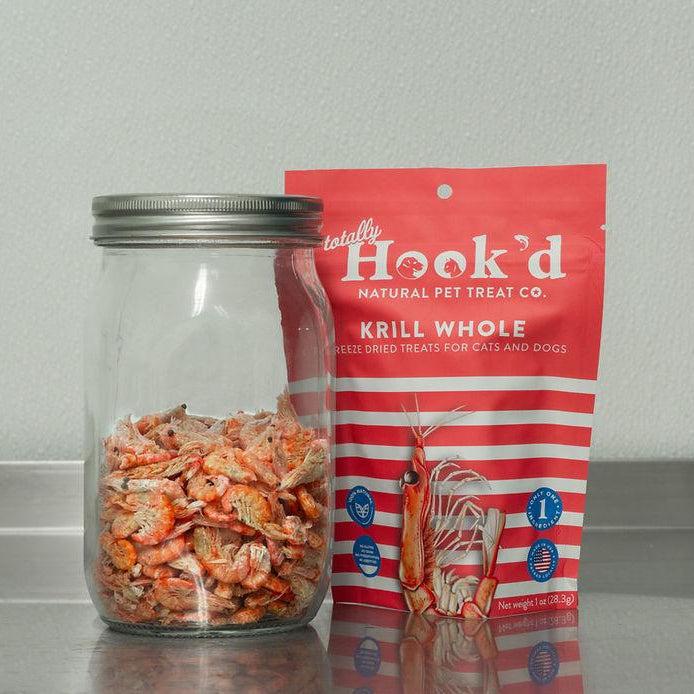 Jar of pet treats next to a red and white package labeled 'Hook'd Krill Whole' on a gray surface.
